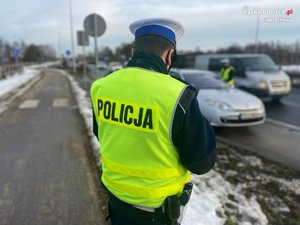 na zdjęciu policjant ruchu drogowego stoi przy drodze - w tle drugi policjant kontroluje kierującego pojazdem