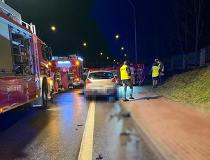 na zdjęciu samochód koloru srebrnego marki Peugeot na miejscu wypadku - w tle widać policjantów i wozy strażackie