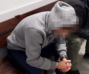Na zdjęciu zatrzymany ubrany w szarą bluzę z zaciągniętym na głowę kapturem siedzi na ławce przed salą rozpraw, obok nieumundurowany policjant.