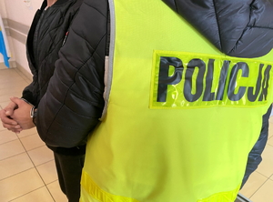 na zdjęciu policjant w kamizelce odblaskowej, za nim zatrzymany w kajdankach założonych na ręce z przodu