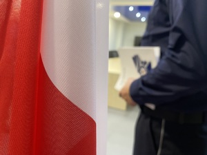 na zdjęciu na pierwszym planie flaga i zbliżenie na teczkę trzymaną w ręce przez policjanta