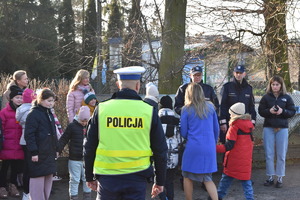 na zdjęciu policjant z ruchu drogowego stoi przodem do uczniów i tłumaczy im zasady bezpieczeństwa przed wejściem na jezdnię
