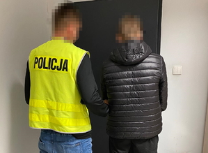 na zdjęciu policjant w kamizelce odblaskowej trzyma pod ramię zatrzymanego mężczyznę