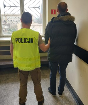 Policjant z zatrzymanym stoi przed zakratowanym oknem