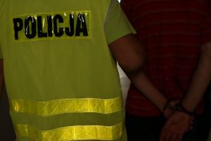 Policjant doprowadza zatrzymanego, który ma kajdanki założone na ręce trzymane z tyłu