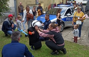 Dziecko przymierza policyjny sprzęt- obok zaparkowany radiowóz  a przy nim uczestnicy pikniku