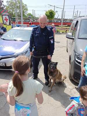 Policjant z psem stoi przy radiowozie, obok przyglądają się dzieci