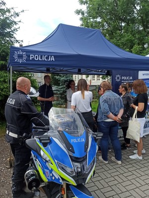 Policjant stoi przy policyjnym motocyklu, a przy policyjnym stanowisku policjanci rozmawiają z uczestnikami targów pracy