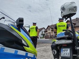 Policjanci stoją przy drodze, za nimi zaparkowany radiowóz i motocykl