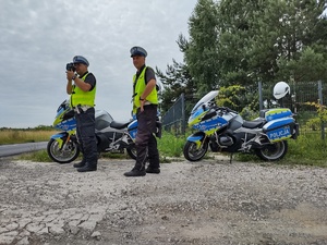 Policjanci stoją przy drodze obok motocykli służbowych i czuwają nad ruchem pojazdów