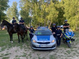 Na zdjęciu widoczne są dwa konie służbowe wraz z policyjnymi jeźdźcami, radiowóz policyjny, a obok niego dwie policjantki i jeden policjant oraz motocykl policyjny wraz z policjantem. W tle drzewa.