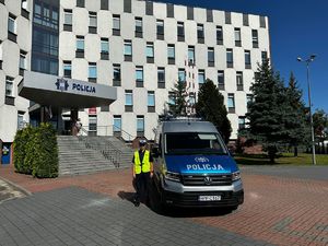 Na pierwszym planie widoczny policjant Wydziału Ruchu Drogowego w kamizelce odblaskowej stojący obok oznakowanego radiowozu typu &quot;furgon&quot;. W tle budynek Komendy Policji.