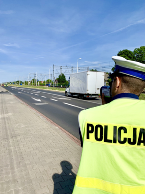 Na pierwszym planie policjant ruchu drogowego widziany od tyłu, w trakcie pomiaru prędkości. W tle droga i poruszające się po niej pojazdy.