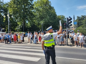 Policjant kieruje ruchem i unosi prawą rękę do góry. Po przejściu dla pieszych przechodzi kolumna pieszych