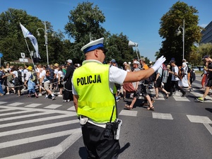 Policjant stoi na drodze i kieruje ruchem pojazdów unosząc prawą rękę do góry. Przy przejściu dla pieszych stoi kolumna pielgrzymów