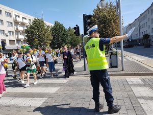 Policjant wyciąga prawą rękę, obok przechodzą przez jezdnię piesi