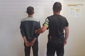 Policjant doprowadza zatrzymanego, który ma kajdanki założone na ręce trzymane z tyłu