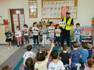 Policjant prowadzi prelekcje na temat bezpieczeństwa z dziećmi, które prezentują tablice z numerami alarmowymi