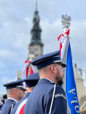 Policjant w mundurze galowy trzyma sztandar- w tle wieża Jasnej Góry