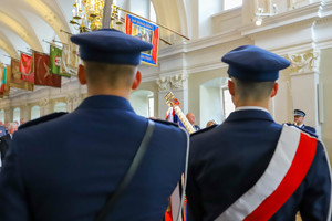 Policjanci odwróceni tyłem stoją ze sztandarem w sali. Jeden z nich ma przewieszoną szarfę z barwami narodowymi