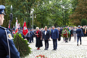 Asysta zaproszonych gości w towarzystwie policjanta idzie z wiązanka kwiatów