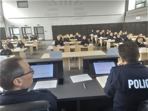 Komendant miejski i jego zastępca siedzą przed monitorem skierowany w stronę siedzących w ławkach policjantów