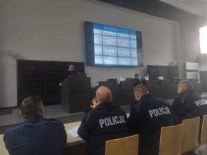 Prelegent przemawia do mikrofonu skierowany w stronę widowni na której zasiadają policjanci w mundurach służbowych