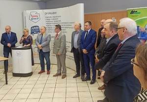 Uczestnicy targów stoją obok siebie w sali- w tle baner reklamowy a przy nim mównica