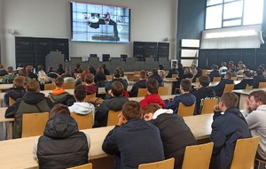 Młodzież siedzi w ławkach w auli i ogląda spot wyświetlany na ekranie