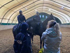 Na pierwszym planie policjant i egzaminator stojący tyłem, przed nimi dwóch jeźdźców policyjnych na koniach służbowych.