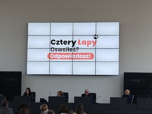 Prelegenci siedzą pod wyświetlaczem na którym wyświetla się hasło kampanii