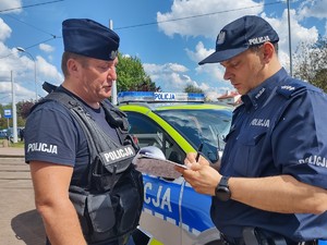 Policjanci rozmawiają przy radiowozie, jeden z nich notuje na kartce