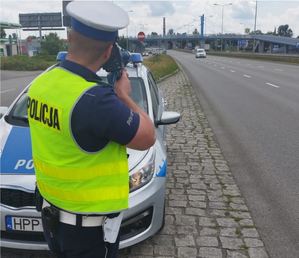 Policjant stoi przy drodze przed radiowozem i kontroluje prędkośc pojazdów