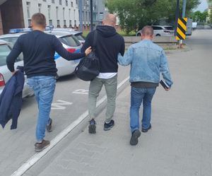 Nieumundurowani policjanci doprowadzają zatrzymanego przez plac komendy obok radiowozów