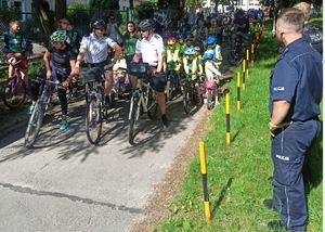 Policjanci na rowerach przygotowują wyścig rowerowy do startu. Za nimi grupa dzieci nz rowerami