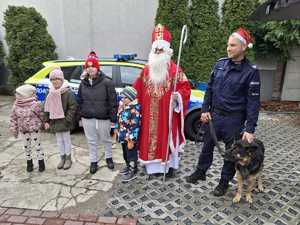 Mikołaj i policjant z psem służbowym pozują do zdjęcia z dziećmi na tle radiowozu