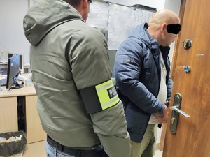 Policjant z opaską na przedramieniu z napisem Policja, doprowadza zatrzymanego do pomieszczenia służbowego