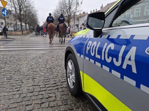 Policyjny radiowóz i policjanci na koniach zabezpieczają przemarsz aleją