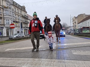 Wolontariusz z dzieckiem przechodzi przez jezdnię, za nim policyjne konie i radiowóz