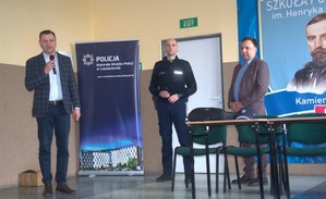 Mężczyzna mówi do mikrofonu, obok stoi policjant i kobieta w sali