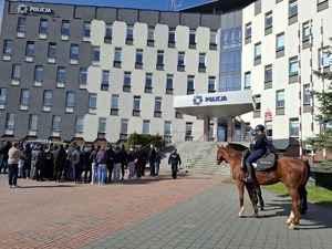 Policyjni jeźdźcy na koniach stoją obok zgromadzonych ludzi przed komendą