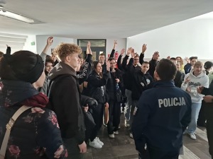 Policjant stoi przed grupą kandydatów, którzy unoszą ręce do góry