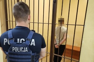 Policjant stoi przed kratami pomieszczenia w którym przebywa zatrzymany w kajdankach