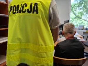 Policjant w pomieszczeniu służbowym stoi przy zatrzymanym, który siedzi na krześle