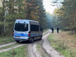 Policjanci idą duktem leśnym przy którym stoi policyjny radiowóz