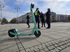 Hulajnoga elektryczna, w tle policyjna kontrola mężczyzny z udziałem dziennikarzy