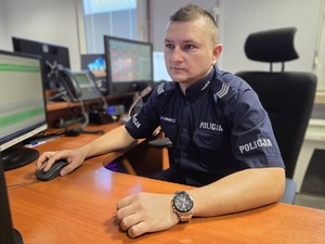Policjant w umundurowaniu służbowym siedzi za biurkiem na stanowisku dyżurnego