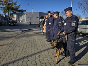 Policjanci z psem służbowym i uczestnicy stoją na placu komendy