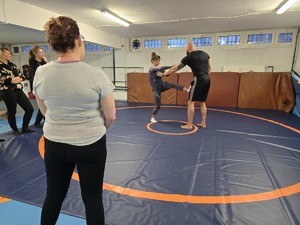 Kobieta wykonuje kopnięcie na macie w stronę instruktora, reszta kobiet przygląda się