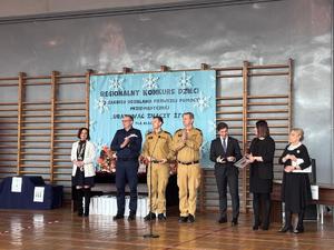 Policjant i strażacy w sali, której odbywa się konkurs stoją przy mikrofonie